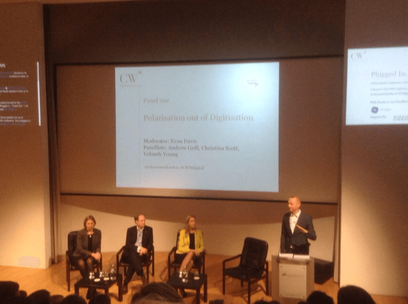 Panel discussion on a stage: five people (four seated, one at a podium) with a presentation slide behind them.